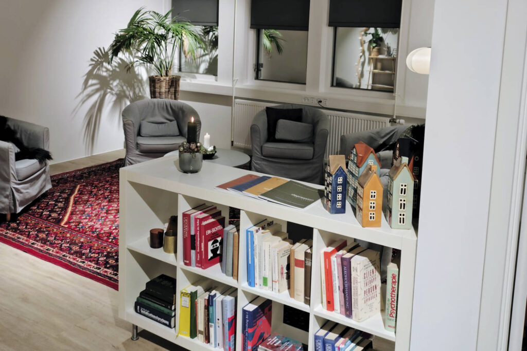 Moderne stueinteriør med hvidt hyldesystem fyldt med bøger, grå lænestole, orientalsk tæppe og grønne planter ved vinduet.