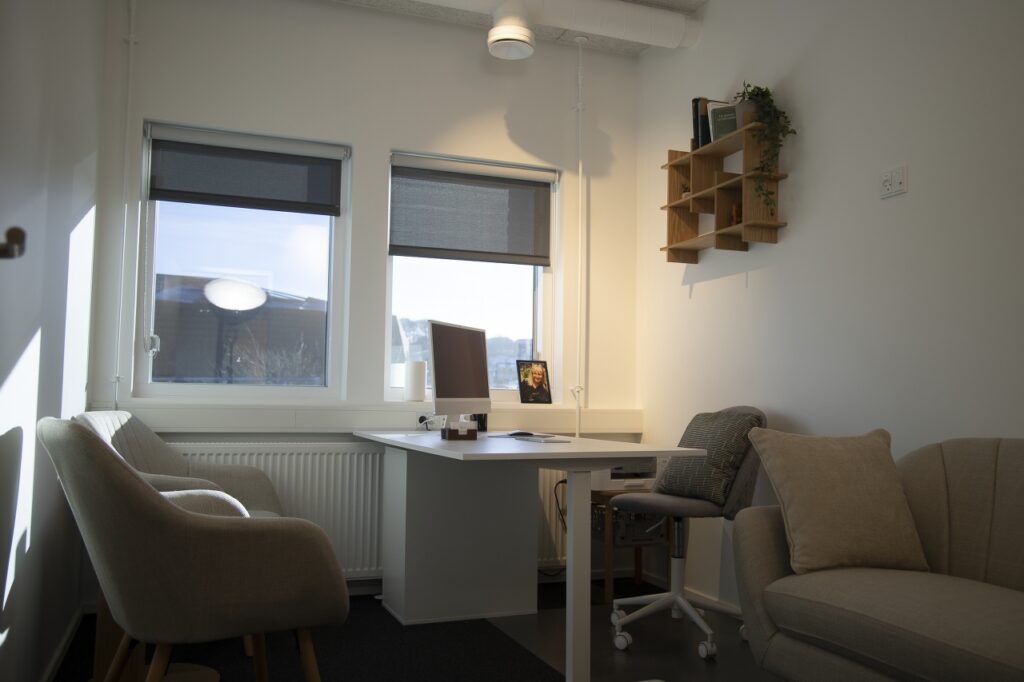Moderne kontorrum med skrivebord, computer, beige lænestol, sofa og store vinduer med udsigt.